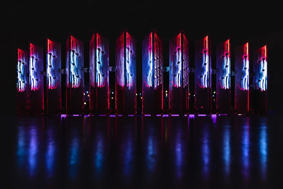 Pou Marama: a row of tall engraved acrylic pou, lit from the side in red and blue so the engraved designs glow, reflected in a dark floor.